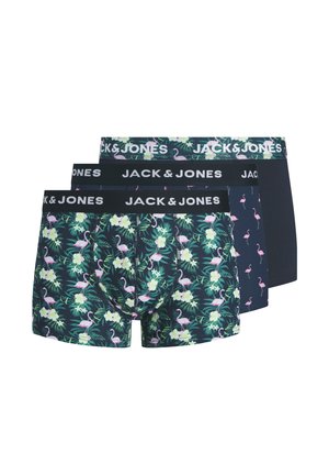 Jonge man staand, draagt donkere boxershorts met roze flamingo- en groene bladerenprint en een "JACK & JONES" tailleband, neutrale achtergrond.