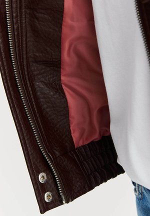 Brązowa skórzana kurtka z fakturowanym wykończeniem, srebrnym zamkiem błyskawicznym oraz kontrastującą różową podszewką, z elastycznymi mankietami na dole.