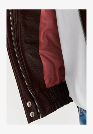 Brązowa skórzana kurtka z fakturowanym wykończeniem, srebrnym zamkiem błyskawicznym oraz kontrastującą różową podszewką, z elastycznymi mankietami na dole.