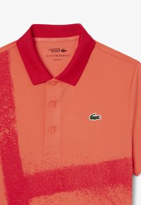 Orange polo som har en röd krage, med en gradientgrafik och en liten grön krokodil-logotyp på vänster sida. Normal passform.