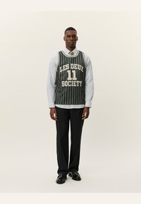 Grøn stribet basketballtrøje med teksten "LES DEUX 11 SOCIETY", båret over en lyseblå skjorte, kombineret med sorte bukser og sko.