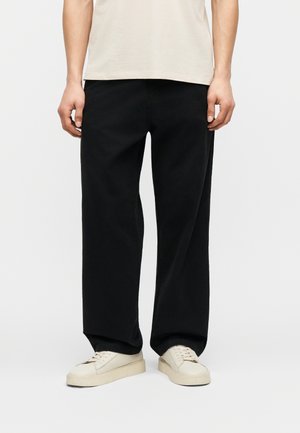 Homme portant un pantalon noir ample, un t-shirt beige et des baskets blanc cassé, debout devant un fond blanc, les mains détendues le long du corps.