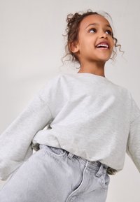 Világosszürke, túlméretezett pulóver széles mandzsettákkal, világos farmer rövidnadrággal párosítva. A modell göndör hajú, felfelé mosolyog. Semleges háttér.