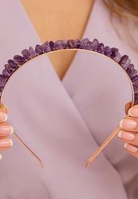 Opaska na głowę w złotym odcieniu ozdobiona nieregularnymi fioletowymi kamieniami ametystu, prezentująca gładki, zakrzywiony kształt i polerowane, błyszczące wykończenie.