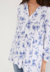 Blusa bianca con stampa floreale blu, dotata di un davanti con bottoni, scollo a V e design a strati con maniche lunghe. Texture in cotone liscia.