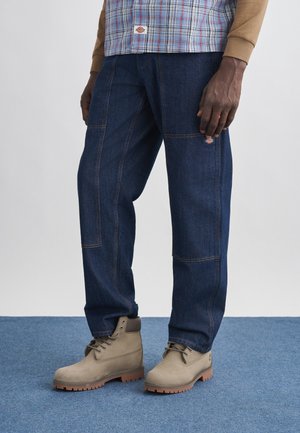 Person iført mørkeblå arbejdsbukser med et lille logo på låret og lysebrune snørestøvler på blåt tæppe.