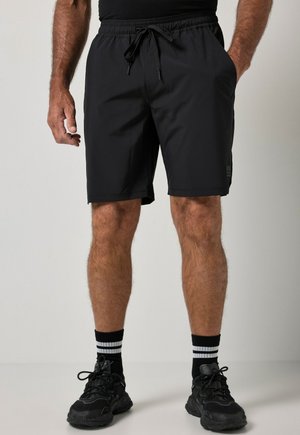 Mand iført sorte snøre shorts, sorte sportssko og sorte sokker med hvide striber, stående mod en ensfarvet baggrund.