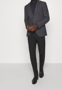 Tmavě kostkovaný blejzr s výstřihy, zapínání na jediný knoflík. Kombinovaný s přiléhavými černými kalhotami a elegantními černými botami. Ušitý na míru.