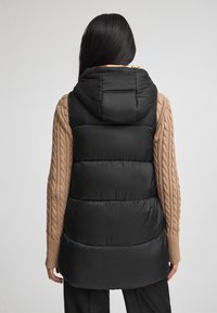 Svart pufferväst med huva, som har en quiltad design. Bärs över en kabelstickad tröja, vilket visar en slät textur och lager-på-lager-look.