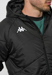 Veste rembourrée noire avec capuche, dotée d'un design matelassé diagonal, d'une fermeture éclair à l'avant et d'un logo brodé blanc sur la poitrine.