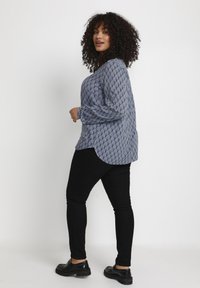 Blå mønstret bluse med en rund kant, lange ærmer, parret med sorte skinny bukser og sorte sko. Krøllet hår, vendt sideværts.