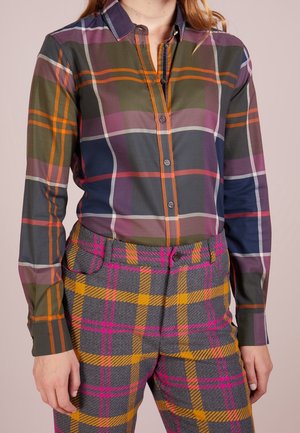 Žena v kostkované košili s dlouhým rukávem v oranžových, fialových a zelených tónech, která je doplněna šedými kalhotami s výraznými růžovými a žlutými kostkami.