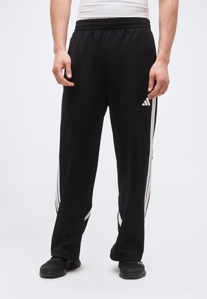 Mężczyzna w czarnych dresowych spodniach Adidas z białymi paskami po bokach i czarnych sneakersach, stojący na jednolitym tle.