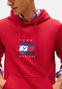 Hombre que lleva una sudadera roja de Tommy Hilfiger con logo y el texto "1985", camiseta de rayas azules y blancas debajo, con las manos en los bolsillos.