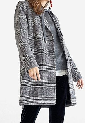 Kobieta ubrana w szary płaszcz w kratę, nałożony na szarą bluzę z kapturem i czarne spodnie, na tle jednolitej białej ściany.