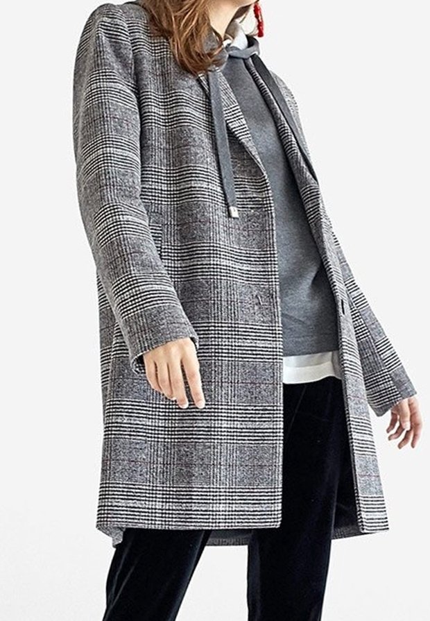 Femme portant un pardessus à carreaux gris superposé à un sweat à capuche gris et un pantalon noir, sur un fond blanc uni.