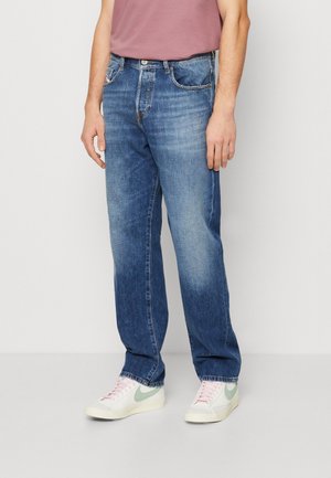 Homme portant un jean bleu délavé droit, un t-shirt rose clair et des baskets blanches avec un swoosh vert clair.