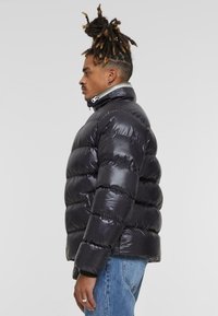 Zwarte pufferjack met een glanzende afwerking, voorzien van horizontaal gestikte delen, een hoge kraag en een zilveren ritsdetail.