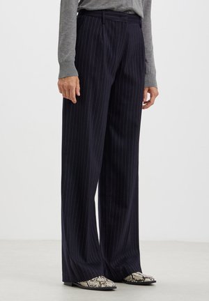 Personne portant un pantalon large bleu marine foncé à fines rayures, un top gris à manches longues et des chaussures pointues à motif peau de serpent noir et blanc.