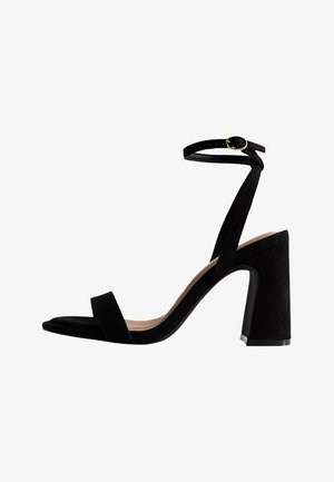 Sandale à talon haut en daim noir avec bride à la cheville et boucle dorée, talon bloc et design à bout ouvert.