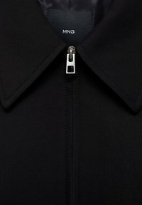 Zwarte zip-up jas met een brede kraag, voorzien van een zilveren rits en een gladde stofstructuur. Het label leest "MNG."