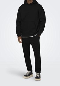 Sudadera de algodón negra con bolsillo tipo canguro, llevada sobre una camiseta blanca, combinada con pantalones de chándal negros a juego y zapatillas negras con suelas blancas.