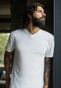 Hvit bomulls v-hals t-skjorte, avslappet passform, båret av en mann med skjegg. Synlige tatoveringer på underarmen. Myk tekstur, ingen mønstre eller aksenter.
