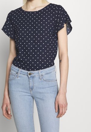 Femme portant un chemisier bleu marine à manches courtes avec des pois blancs et un jean taille haute bleu clair.