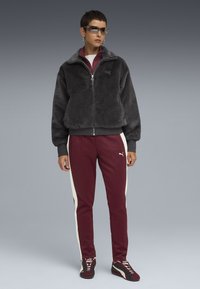 Person standing wearing black fuzzy zip-up jacket, maroon track pants with white side stripes, matching sneakers, and tinted sunglasses.