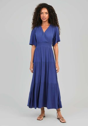 Femme aux cheveux bouclés portant une longue robe bleue à manches courtes avec un col en V et des sandales plates noires à brides, debout devant un fond gris.