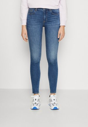 Personne portant un jean skinny bleu, des baskets blanches avec des détails bleus et un haut à manches longues rose clair contre un mur blanc.
