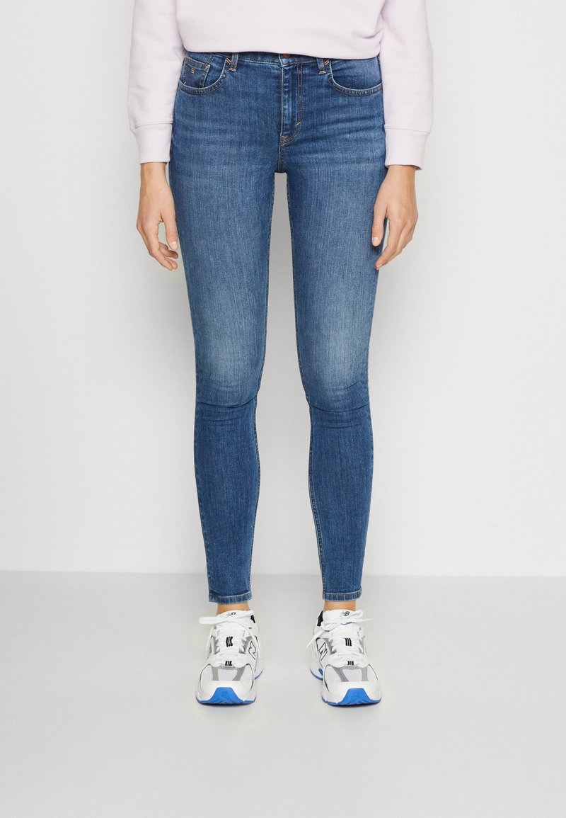 Personne portant un jean skinny bleu, des baskets blanches avec des détails bleus et un haut à manches longues rose clair contre un mur blanc.