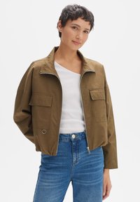 Olivgrüne kurz geschnittene Jacke mit Reißverschluss, großen Klappentaschen und Stehkragen, kombiniert mit einem weißen Oberteil und blauen Jeans.