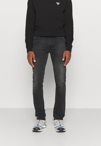 Man wearing black Barbour sweatshirt, dark gray jeans, and gray and white athletic sneakers standing against a plain background.