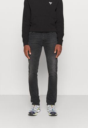 Mies, joka pukeutuu mustaan Barbour-sweatshirthiin, tummanharmaisiin farkkuihin ja harmaisiin sekä valkoisiin urheilukenkään, seisoo yksinkertaista taustaa vasten.