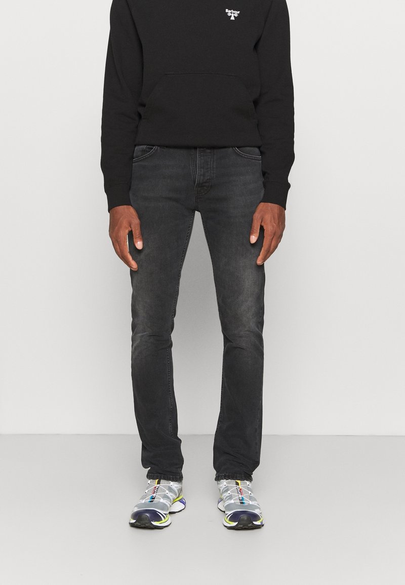 Man wearing black Barbour sweatshirt, dark gray jeans, and gray and white athletic sneakers standing against a plain background.