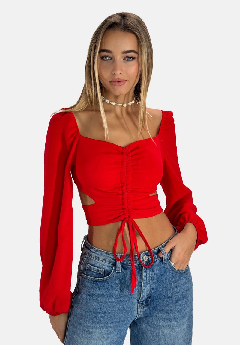 Top corto rosso con scollatura quadrata, fronte arricciato, maniche lunghe a sbuffo e tagli laterali. Abbinato a jeans in denim a vita alta.