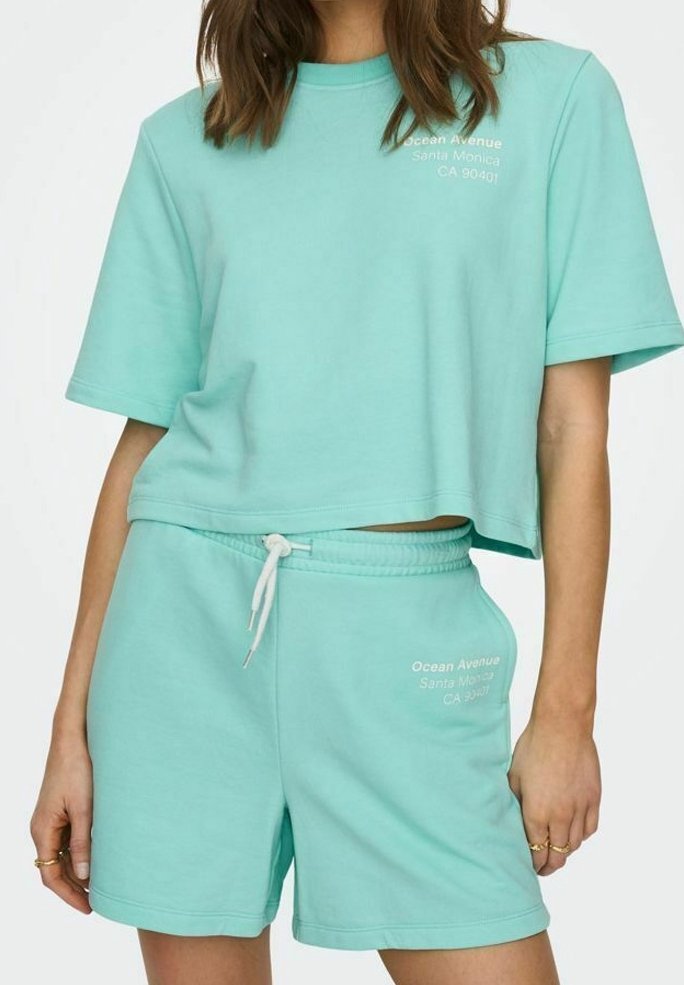 T-shirt turquoise coupé et shorts assortis en tissu doux. Le T-shirt présente un texte blanc ; les shorts ont une taille élastique avec cordon de serrage.