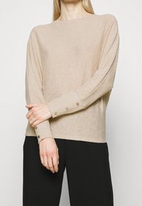 Beiger Strickpullover mit lockerem Schnitt, rundem Ausschnitt und langen Ärmeln. Knöpfe in kontrastierenden Farben an den Bündchen. Weiche Textur.