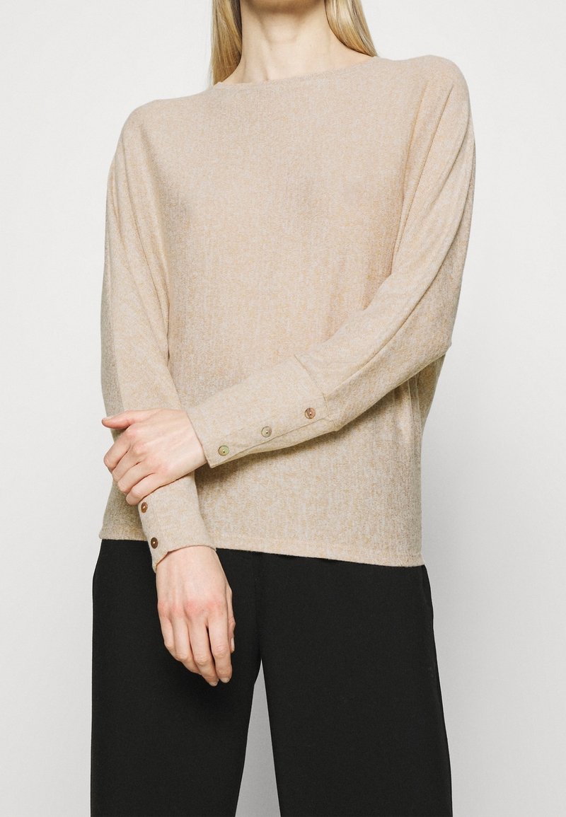 Beiger Strickpullover mit lockerem Schnitt, rundem Ausschnitt und langen Ärmeln. Knöpfe in kontrastierenden Farben an den Bündchen. Weiche Textur.