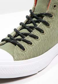 Sneaker alte in tessuto verde con lacci neri, punta bianca e tessuto decorato. Presenta occhielli rinforzati e texture sottili.