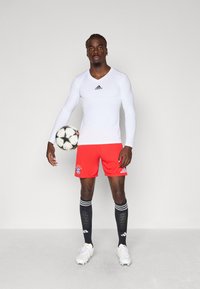 Vit långärmad idrottströja med svart adidas-logotyp, röda shorts med Bayern Münchens emblem, svarta knähöga strumpor och vita fotbollsskor.