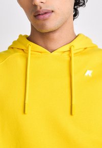 Gros plan d'une personne portant un sweat à capuche jaune vif avec cordons et un petit logo blanc sur la poitrine.