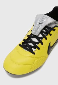 Zapato de fútbol Nike de cuero amarillo con cordones negros, panel lateral perforado, lengüeta plateada y logo de swoosh negro sobre fondo blanco.