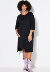 Vestido negro de longitud a la rodilla con mangas tres cuartos, que presenta un diseño de parches texturizados y bolsillos laterales, combinado con botas de vaquero metálicas moradas.
