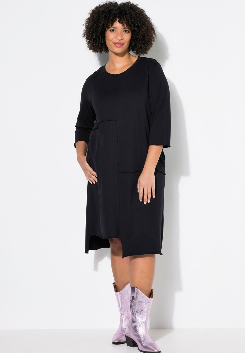Vestido negro de longitud a la rodilla con mangas tres cuartos, que presenta un diseño de parches texturizados y bolsillos laterales, combinado con botas de vaquero metálicas moradas.