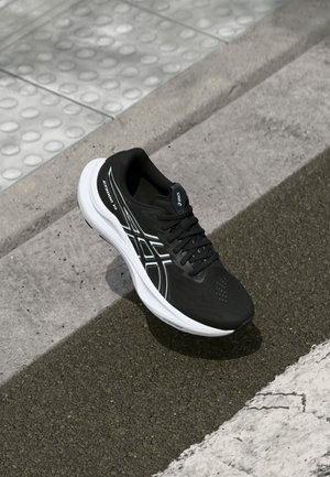 Chaussure de sport noire avec une tige texturée, semelle blanche et accents réfléchissants. Caractéristiques d'un design à lacets et d'un bout perforé pour une meilleure respirabilité.