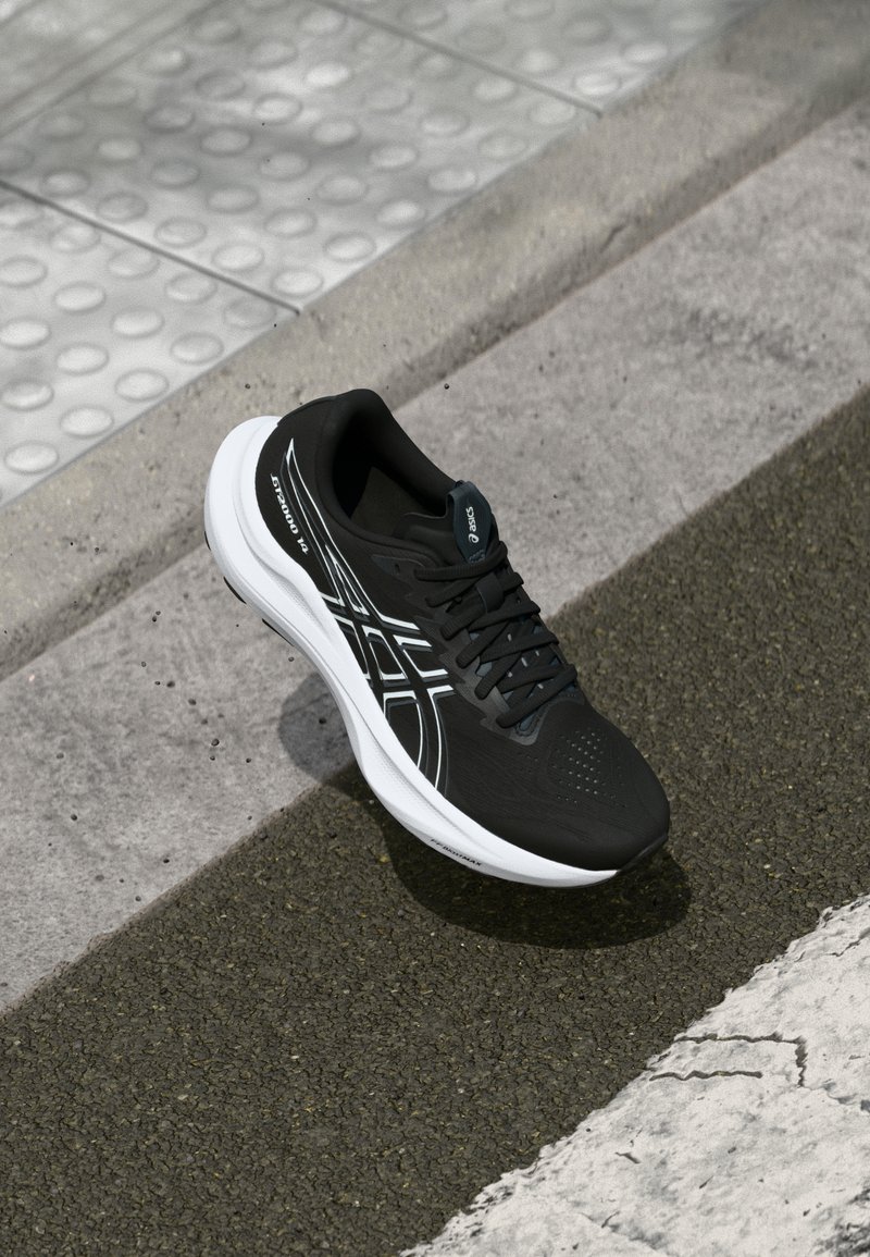 Chaussure de sport noire avec une tige texturée, semelle blanche et accents réfléchissants. Caractéristiques d'un design à lacets et d'un bout perforé pour une meilleure respirabilité.