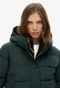 Mørkegrønne pufferjakke med hætte, med en frontlynlås, trykknapper og side lommer. Let tekstureret stof med synlige quiltede sektioner.