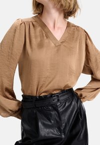 Blouse à manches longues et col en V en satin marron clair avec des détails plissés aux épaules, associée à un pantalon en cuir noir taille haute et une ceinture assortie.